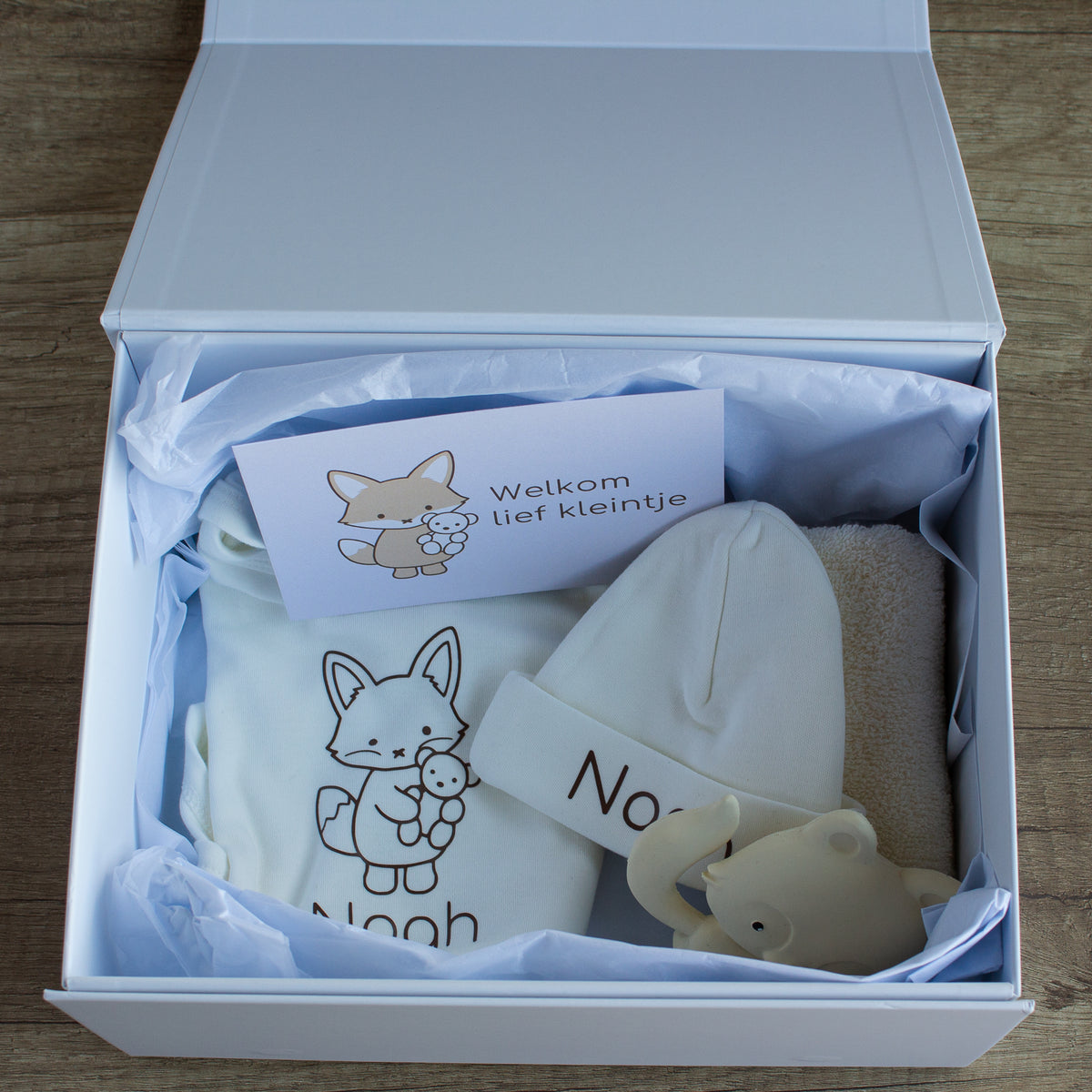 Geopende witte luxe baby giftbox met ecru rompertje en mutsje, gepersonaliseerd met de naam Noah en een vosje, zacht ecru spuugdoekje, crèmekleurig vosje als bijtspeeltje en een kaart met de tekst ‘Welkom lief kleintje’, verpakt in wit vloeipapier.