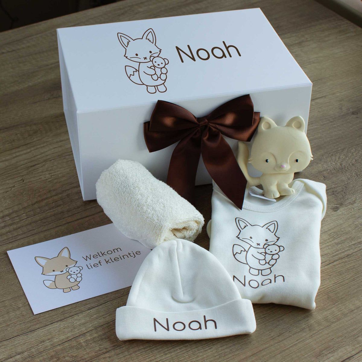 Luxe babycadeauset met vosje in ecru, met een gepersonaliseerde romper en muts met de naam Noah, een crèmekleurig vosje als bijtspeeltje, een ecru spuugdoekje en een kaart met de tekst ‘Welkom lief kleintje’. Achter de producten staat de witte luxe cadeaudoos met vosillustratie en naam Noah, afgewerkt met een chocoladebruine satijnen strik.