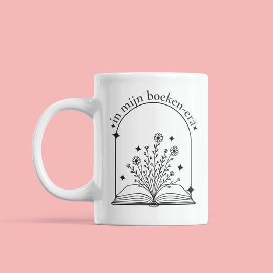 Witte mok met minimalistische illustratie van bloemenboek en tekst “in mijn boeken-era” op roze achtergrond