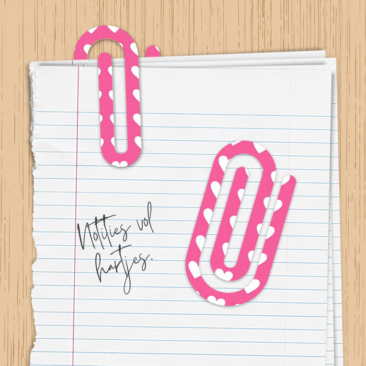 Roze jumbo paperclip met wit hartjespatroon op een notitieblad met handgeschreven tekst "Notities vol hartjes", tegen een houten achtergrond.