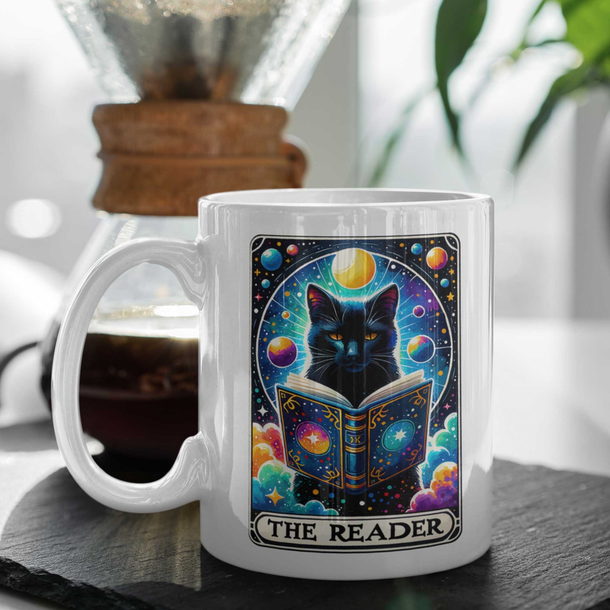 Witte mok met zwarte kat die in een magisch boek leest, tarotstijl design, tekst The Reader, naast koffiezetapparaat