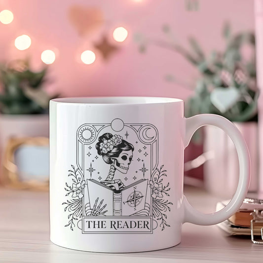 Witte mok met zwart-witte tarotstijl illustratie skelet dat leest, bloemen en symbolen, tekst The Reader, op roze achtergrond met lichtjes