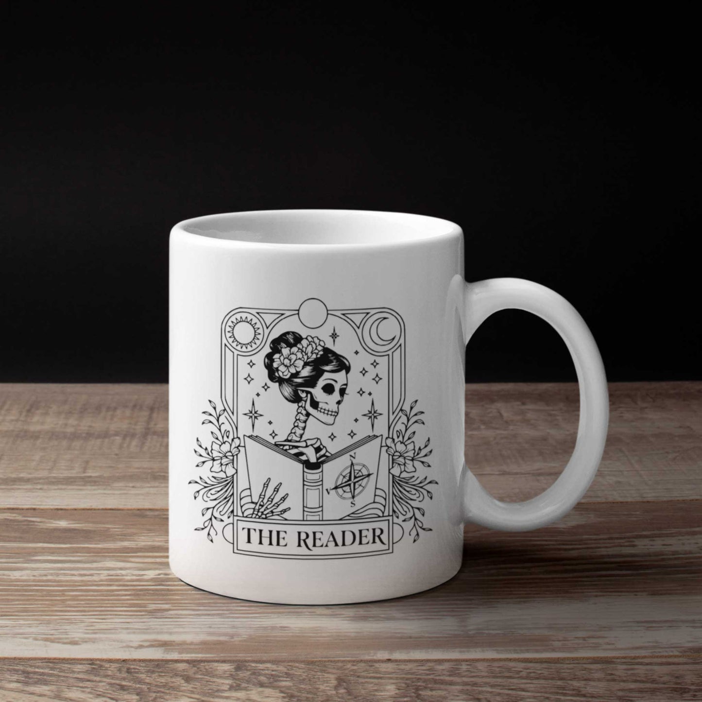 Witte mok met tarotillustratie van skelet met bloemen in haar dat een boek leest, tekst The Reader, op houten tafel