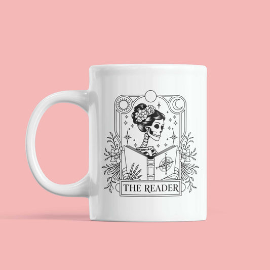 Keramische mok met gothic tarotdesign van skelet en boek, tekst The Reader, witte mok op roze achtergrond