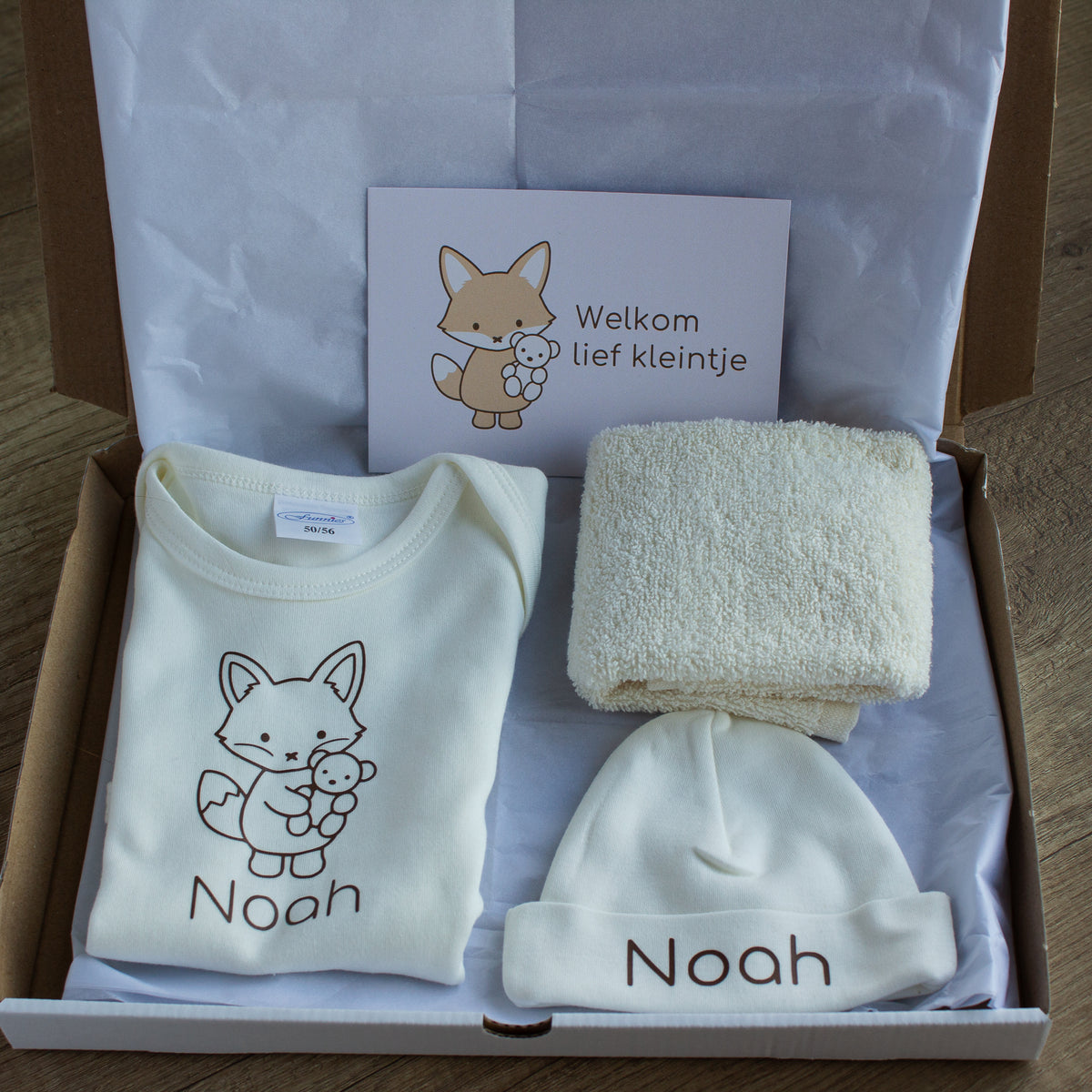 Foto 2 – open doos
Open verzenddoos met ecru newborn cadeauset, bestaande uit gepersonaliseerd rompertje en mutsje met naam Noah, spuugdoekje en cadeaukaartje met vosje.