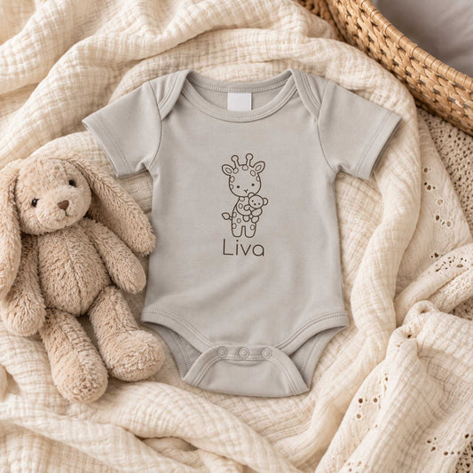Sandstone Grey babyromper met gepersonaliseerde giraf en naam, liggend op zachte lichte stoffen naast een beige knuffel