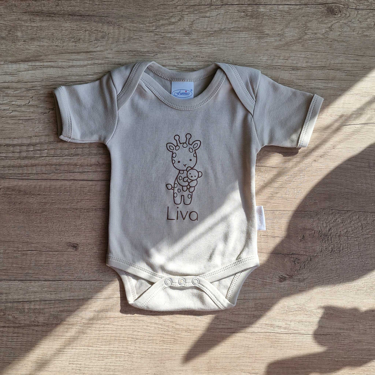 Sandstone Grey babyromper met giraf en gepersonaliseerde naam, liggend op een houten tafel in natuurlijk zonlicht