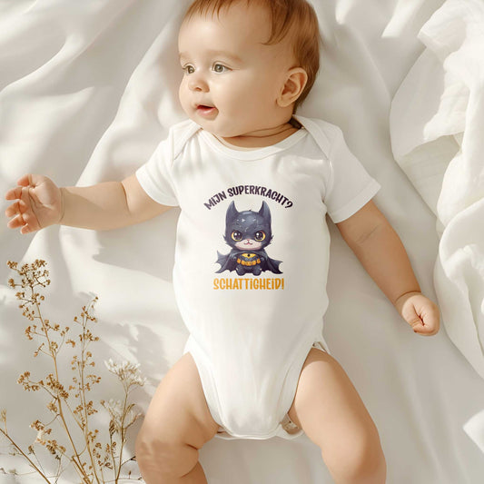 Baby ligt op wit bed in rompertje met superheld-kat en tekst “Mijn superkracht? Schattigheid!” – schattig en neutraal kraamcadeau