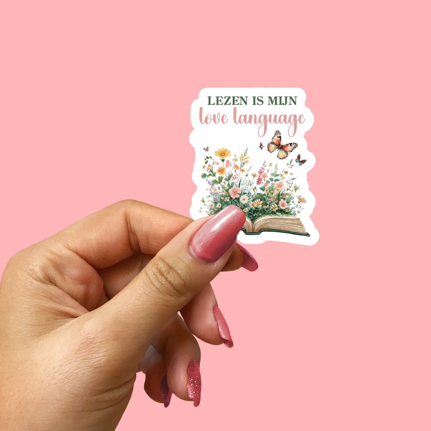 Hand houdt kleine sticker vast met open boek, bloemen en vlinder, tekst Lezen is mijn love language, roze achtergrond