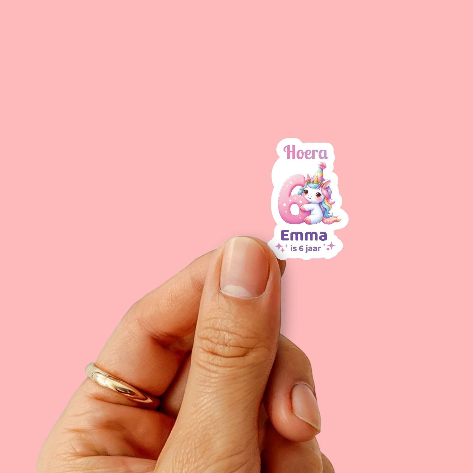 Hand houdt één unicorn sticker met tekst ‘Hoera Emma is 6 jaar’ tegen roze achtergrond
