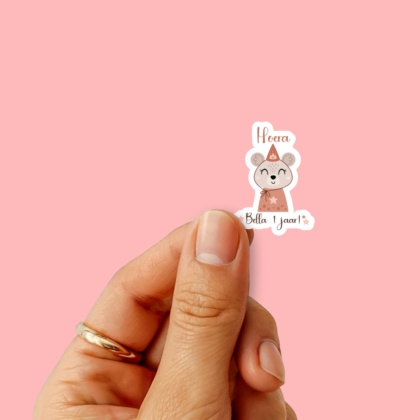 Vinger houdt een muis-sticker vast met tekst ‘Hoera Bella 1 jaar!’ tegen een roze achtergrond