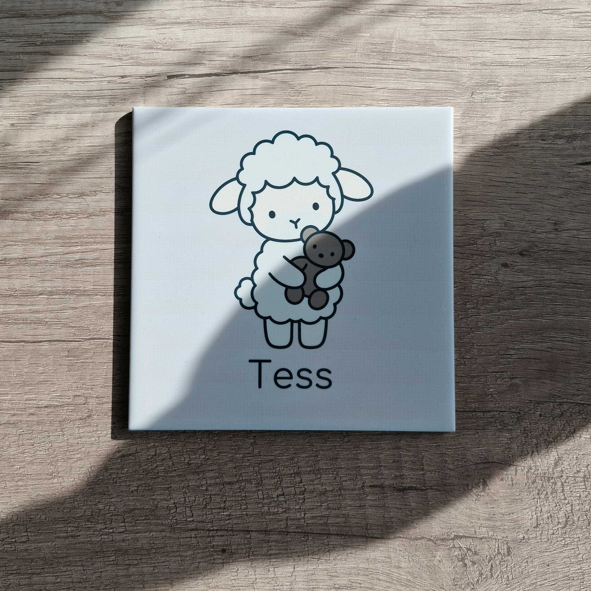 Keramisch tegeltje met een lammetje dat een beertje vasthoudt en de naam Tess, liggend op een houten tafel in zonlicht en schaduw.