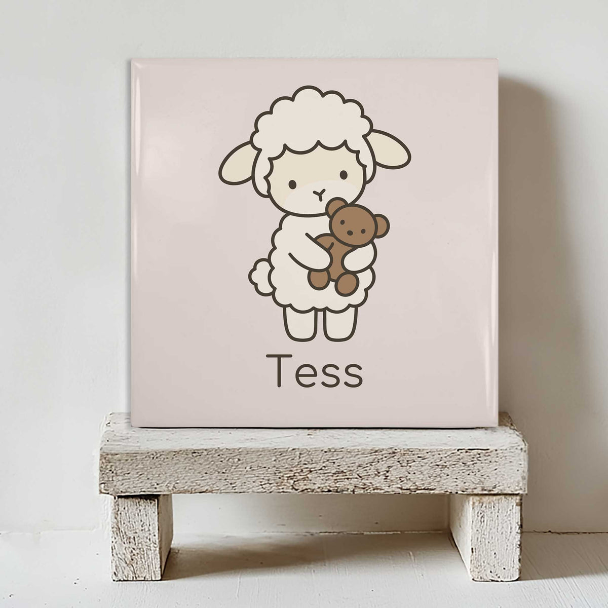 Vierkant keramisch tegeltje met een lammetje dat een beertje vasthoudt en de naam Tess, staand op een wit houten krukje.