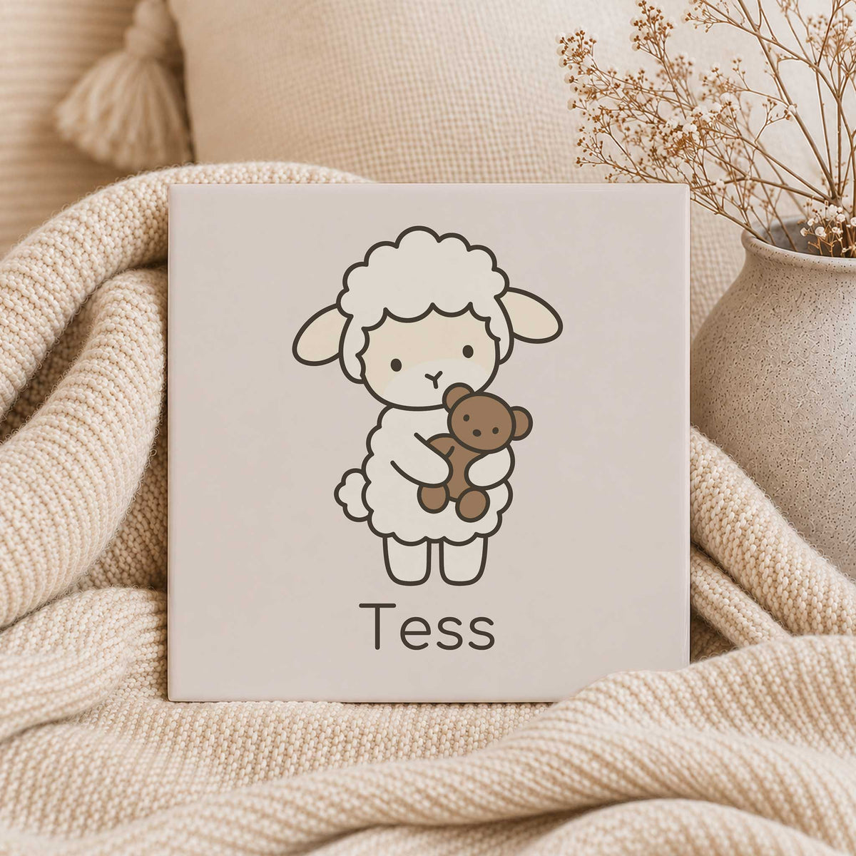 Keramisch tegeltje met een lammetje dat een beertje vasthoudt en de naam Tess, tussen een zachte gebreide deken en een beige vaas.