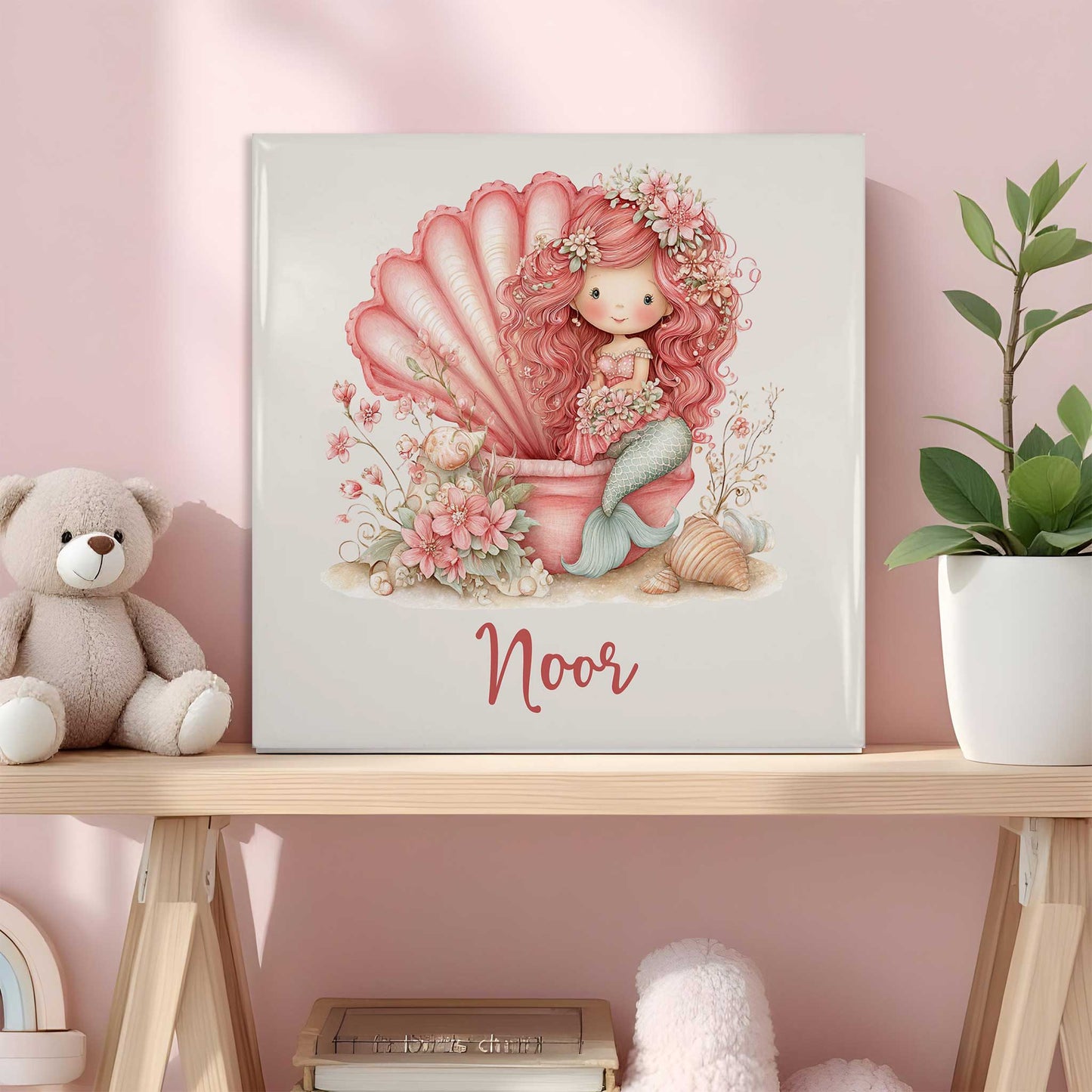 Keramisch tegeltje met de naam Noor. Op het ontwerp staat een roze zeemeermin met lang krullend haar die in een grote roze schelp zit. Rondom haar staan bloemen, kleine schelpen en zachte pastelaccenten. Het tegeltje staat op een houten plank in een roze kinderkamer naast een knuffelbeer en een plant.