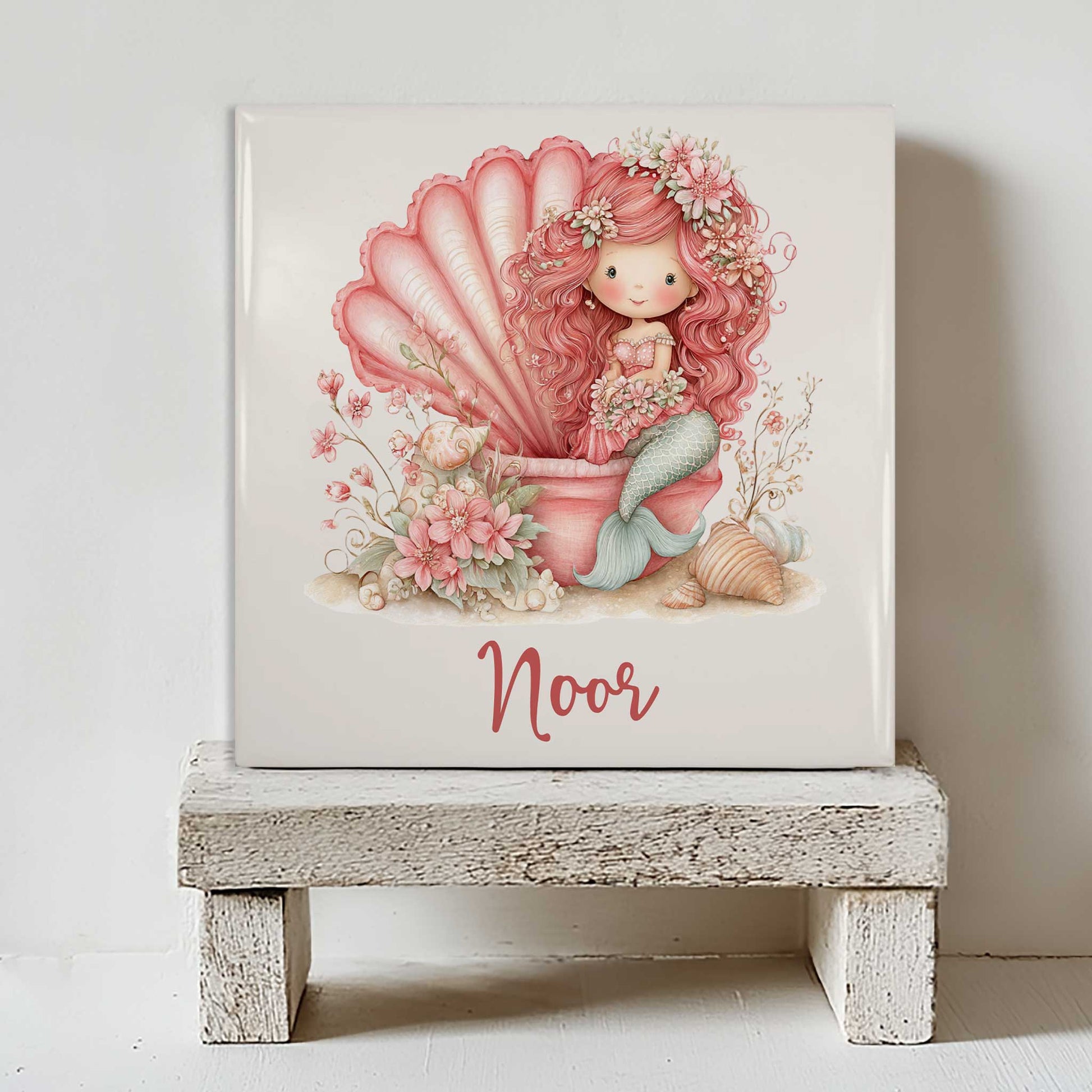 Gepersonaliseerd keramisch tegeltje met een roze zeemeermin in een grote schelp. Het tegeltje staat op een wit houten krukje tegen een lichte muur. Onder de illustratie staat de naam Noor.