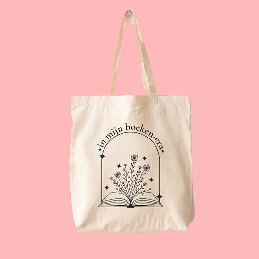 Minimalistische tote bag met zwarte opdruk van bloemen die uit een boek groeien en de tekst “in mijn boeken-era”, op een roze achtergrond.