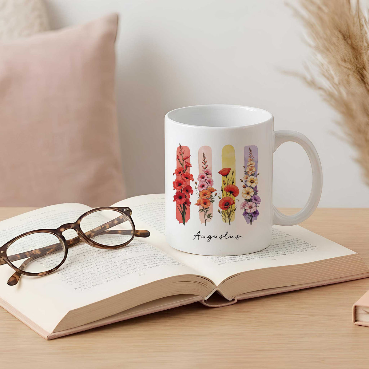 Witte mok met de geboortebloemen gladiool en klaproos en de tekst Augustus, geplaatst op een open boek met een bril op een houten tafel.