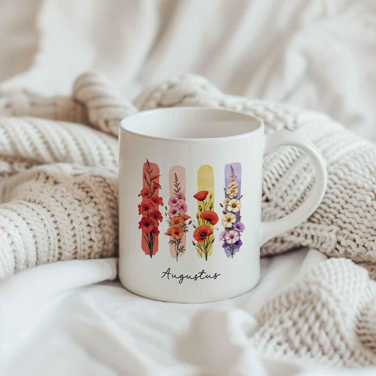 Witte mok met de geboortebloemen gladiool en klaproos en de tekst Augustus, geplaatst tussen beige gebreide dekens.