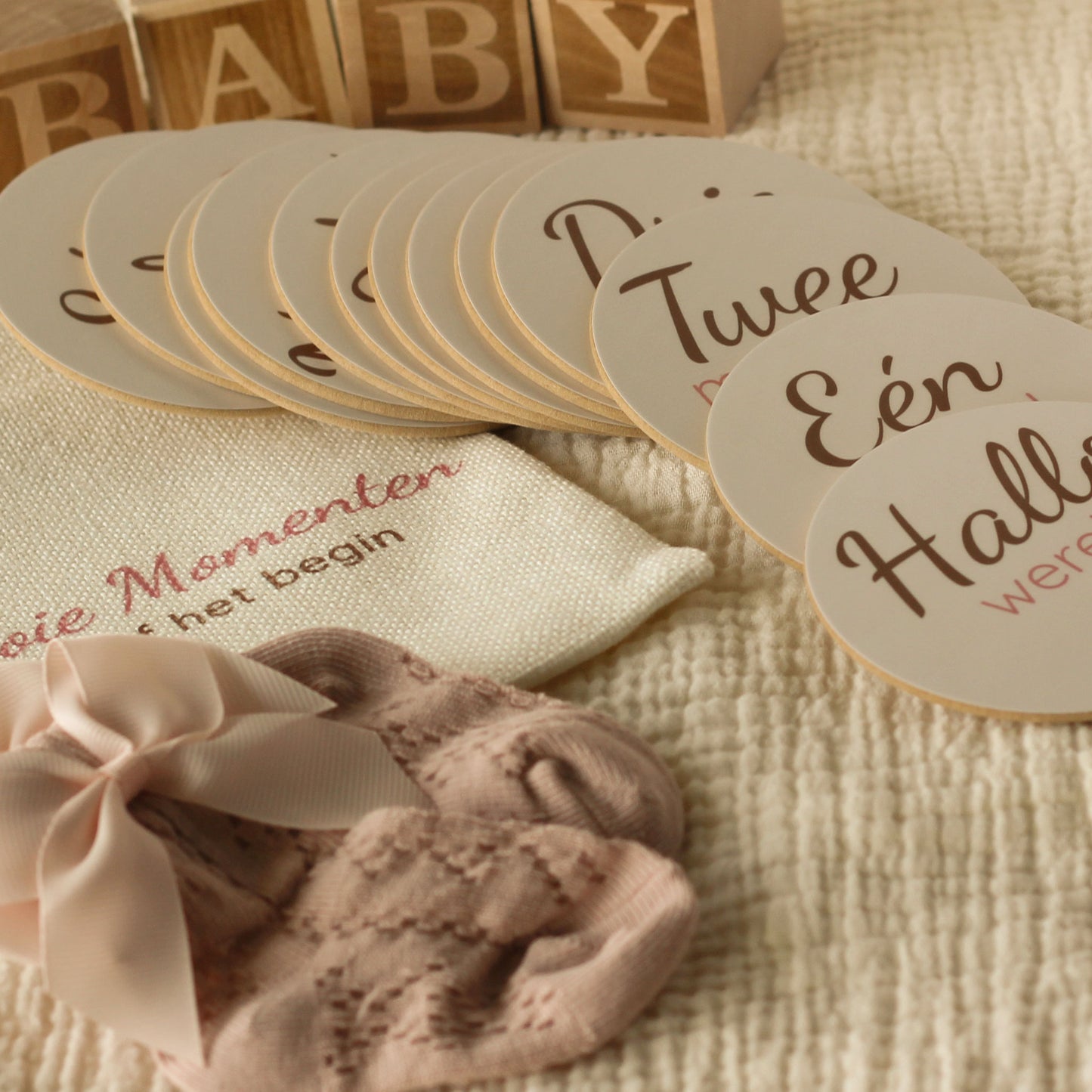 Baby mijlpaalschijfjes gedeeltelijk uitgestald naast een linnen cadeauzakje met tekst, roze babysokjes met strik en houten BABY blokken op een zachte beige hydrofiele ondergrond in warme neutrale tinten