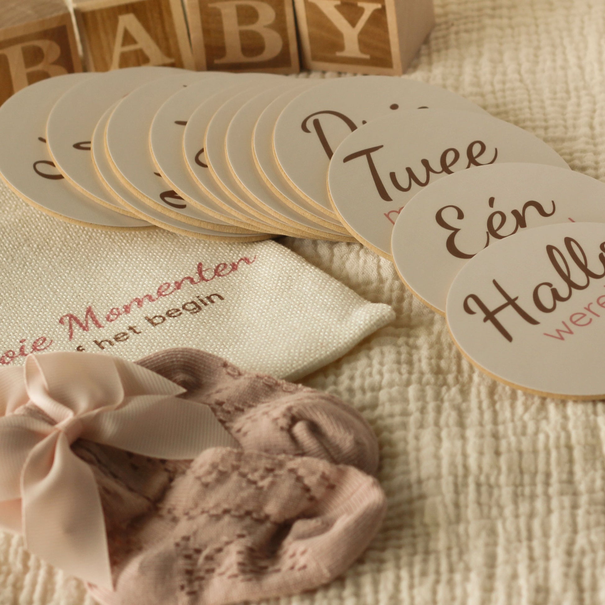 Baby mijlpaalschijfjes gedeeltelijk uitgestald naast een linnen cadeauzakje met tekst, roze babysokjes met strik en houten BABY blokken op een zachte beige hydrofiele ondergrond in warme neutrale tinten