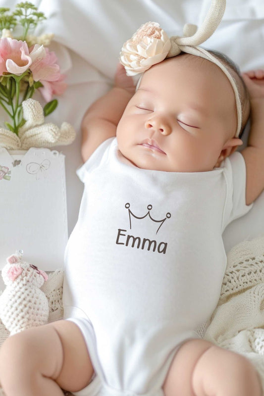 Baby draagt een wit gepersonaliseerd rompertje met naam Emma en minimalistisch kroontje terwijl het kindje op een lichte ondergrond ligt.