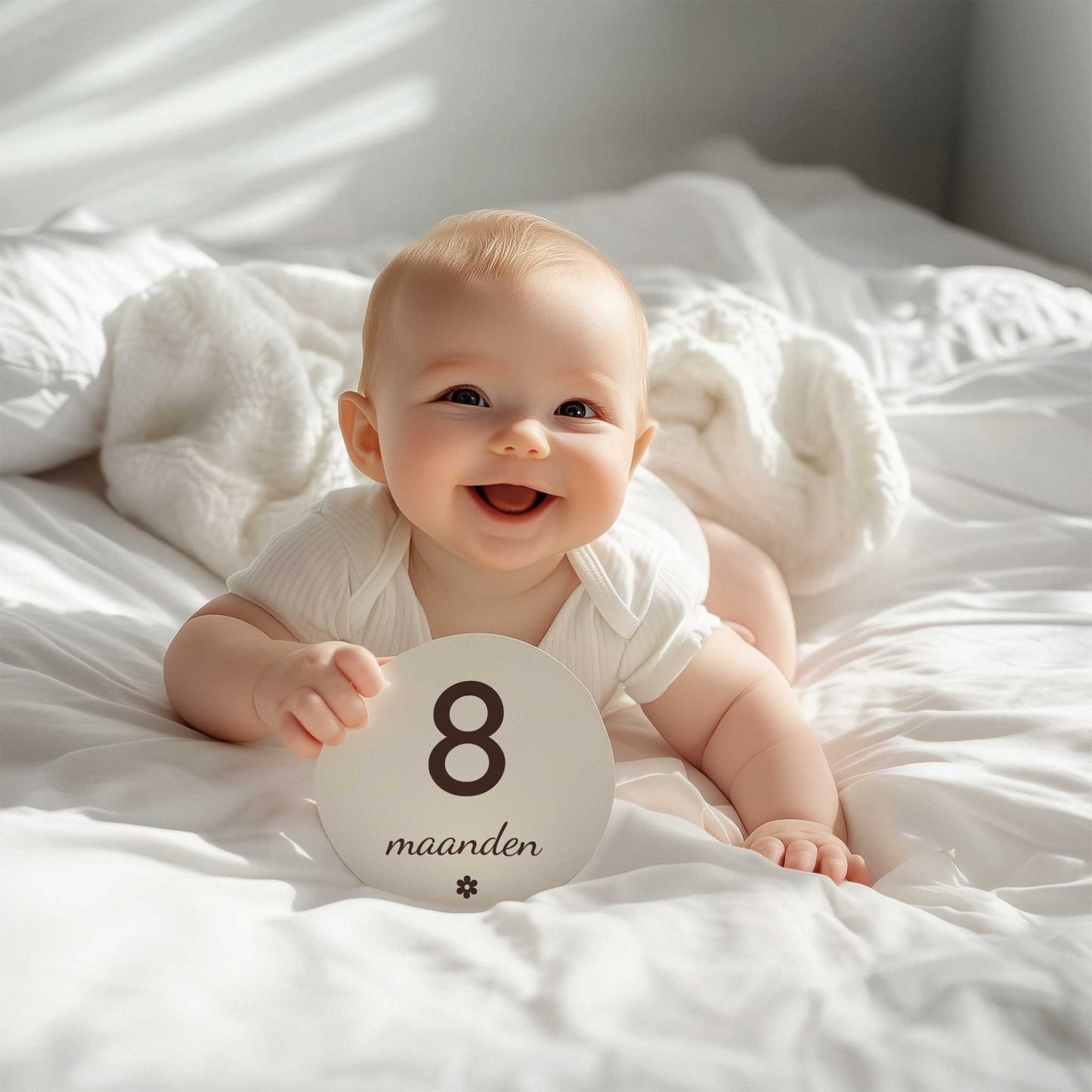 Lachende baby ligt op een wit bed en houdt een ronde mijlpaalschijf met de tekst “8 maanden” vast.
