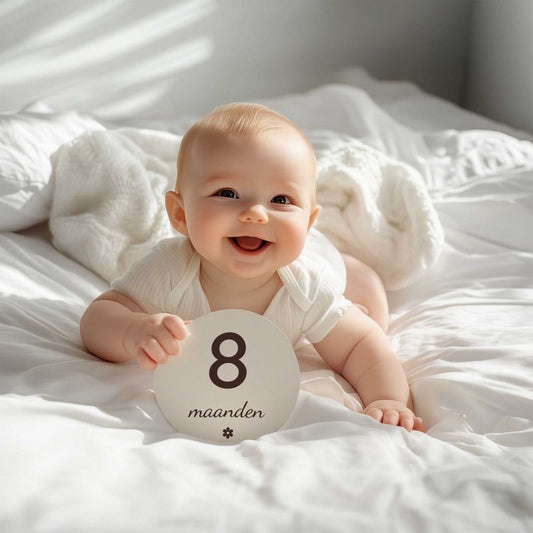 Lachende baby ligt op een wit bed en houdt een ronde mijlpaalschijf met de tekst “8 maanden” vast.