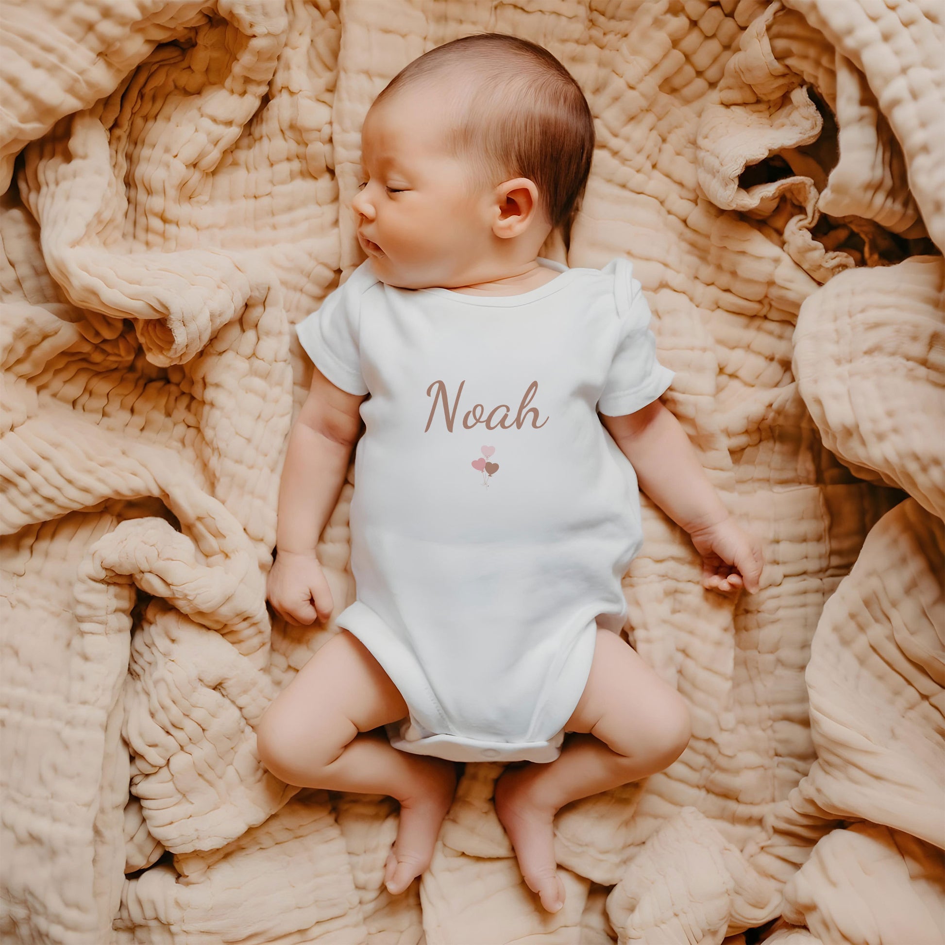 Baby liggend op een zachte beige deken draagt een wit rompertje met de naam Noah en een klein blush ballonnen ontwerp op de borst.