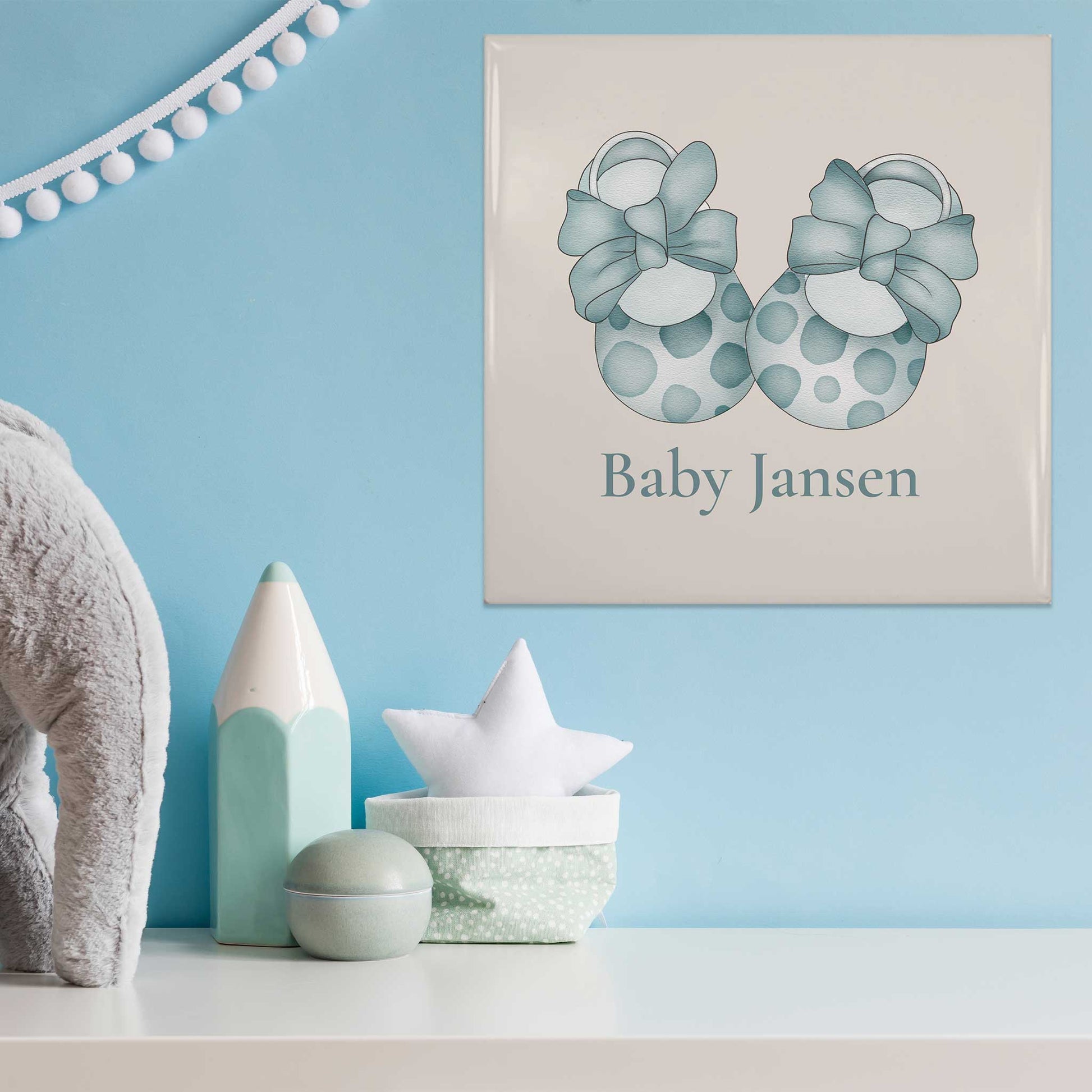 Gepersonaliseerd keramisch baby tegeltje met blauwe babyschoentjes en de naam Baby Jansen in een blauwe babykamer met decoratie op een witte plank.