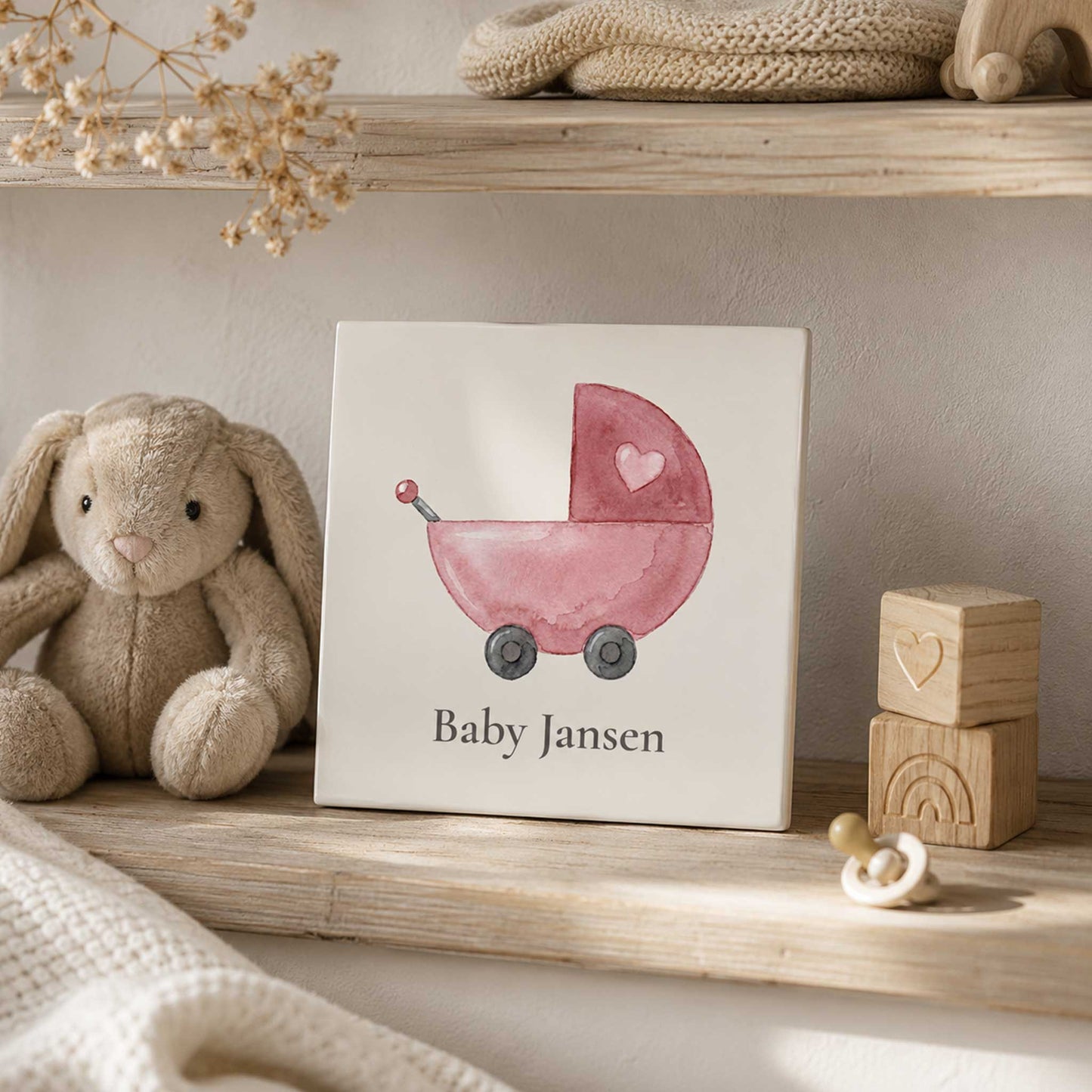 Keramisch babytegeltje met roze kinderwagen illustratie gestyled in een warme babykamer met knuffelkonijn en houten accessoires