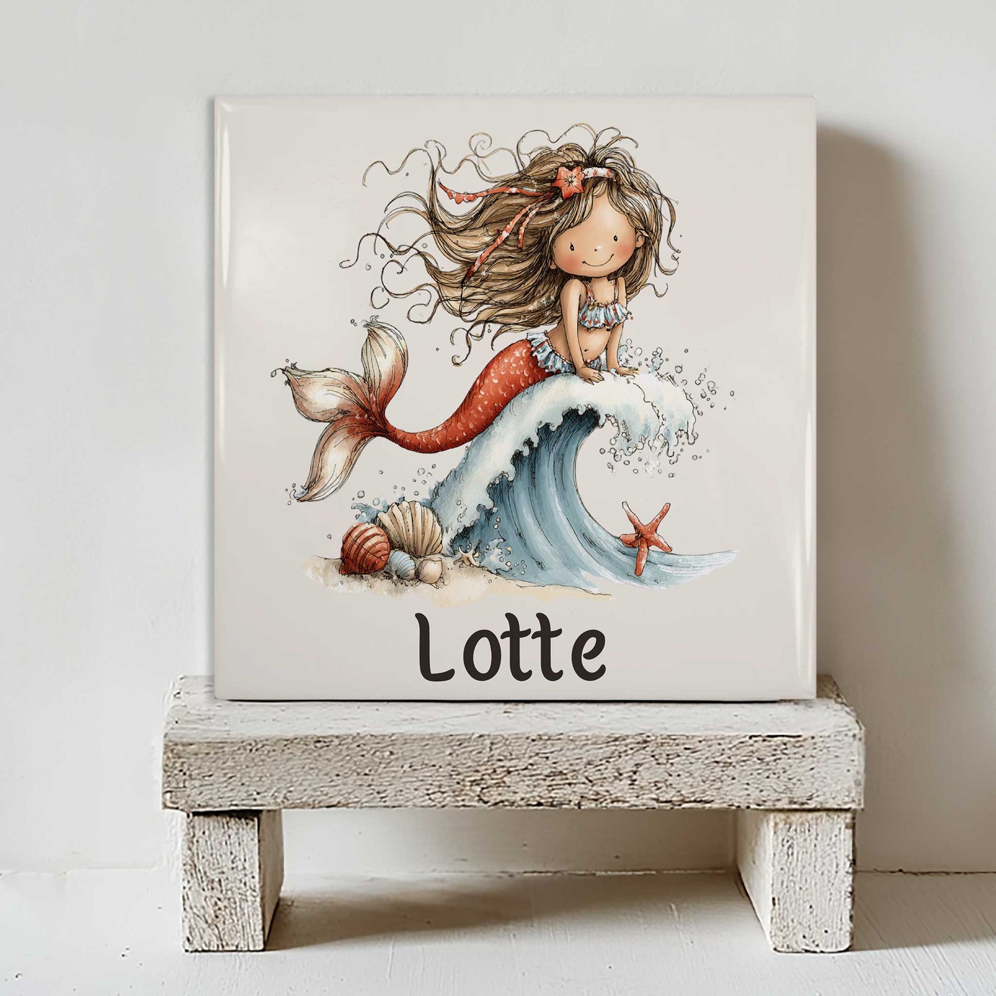 Keramisch tegeltje met een zeemeermin op een schuimende golf en de naam Lotte, geplaatst op een wit houten krukje tegen een lichte muur.