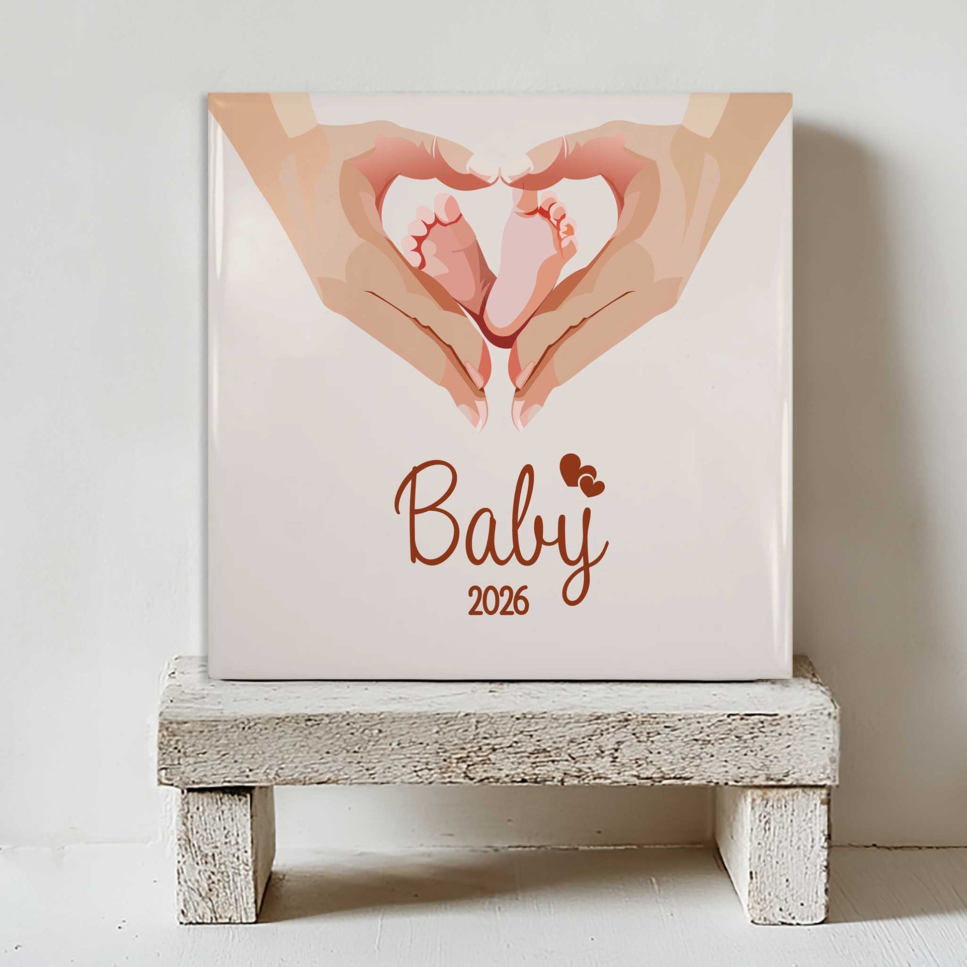 Keramisch babytegeltje met babyvoetjes illustratie en de tekst Baby 2026 op klein wit houten krukje tegen lichte muur