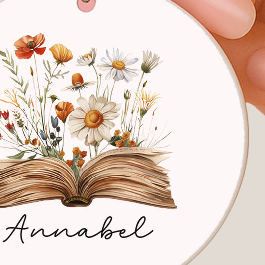 Close-up van een ronde sleutelhanger met een open boek waar bloemen uit groeien en de naam Annabel.