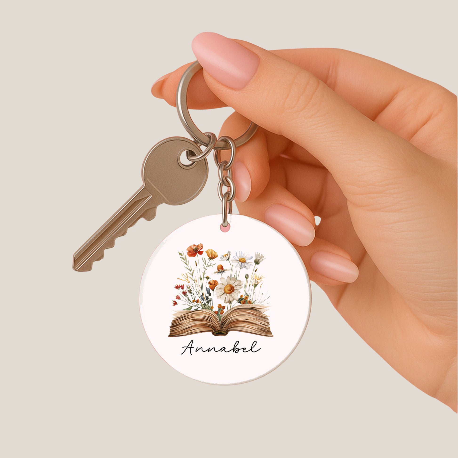 Hand houdt een ronde sleutelhanger vast met een open boek waar bloemen uit groeien en de naam Annabel, samen met een sleutel.