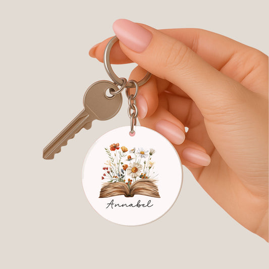 Hand houdt een ronde sleutelhanger vast met een open boek waar bloemen uit groeien en de naam Annabel, samen met een sleutel.