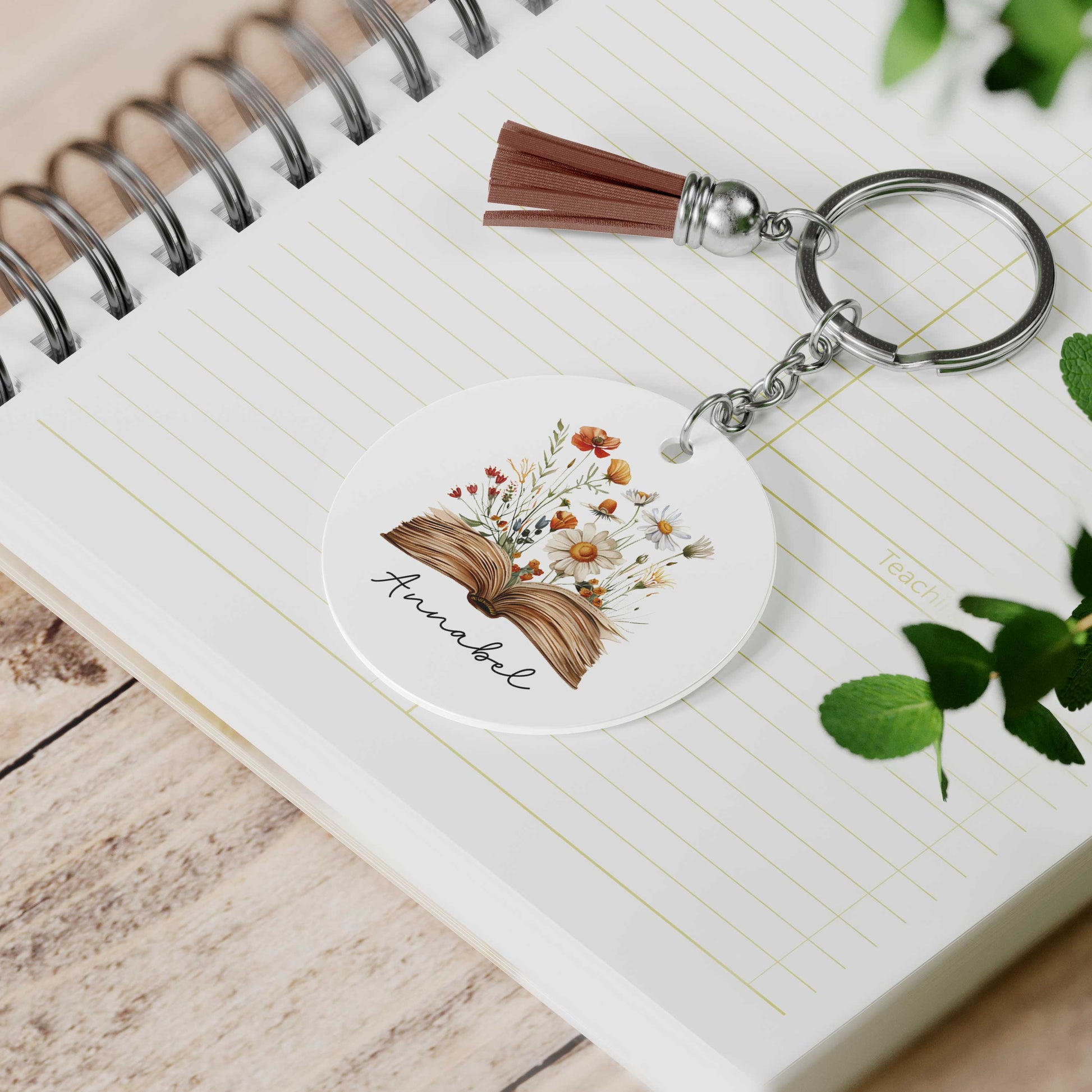 Ronde sleutelhanger met een open boek waar bloemen uit groeien en de naam Annabel, liggend op een open notitieboek naast een bruine tassel.