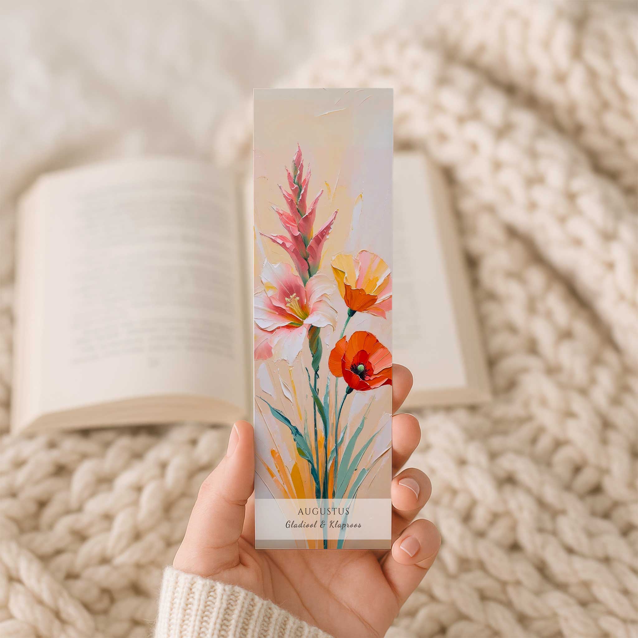 Hand houdt een boekenlegger met de geboortebloemen van augustus – gladiool en klaproos – voor een open boek op een beige gebreide deken. De kunstzinnige illustratie toont kleurrijke bloemen in roze, wit, oranje en rood met onderaan de tekst 'Augustus'.