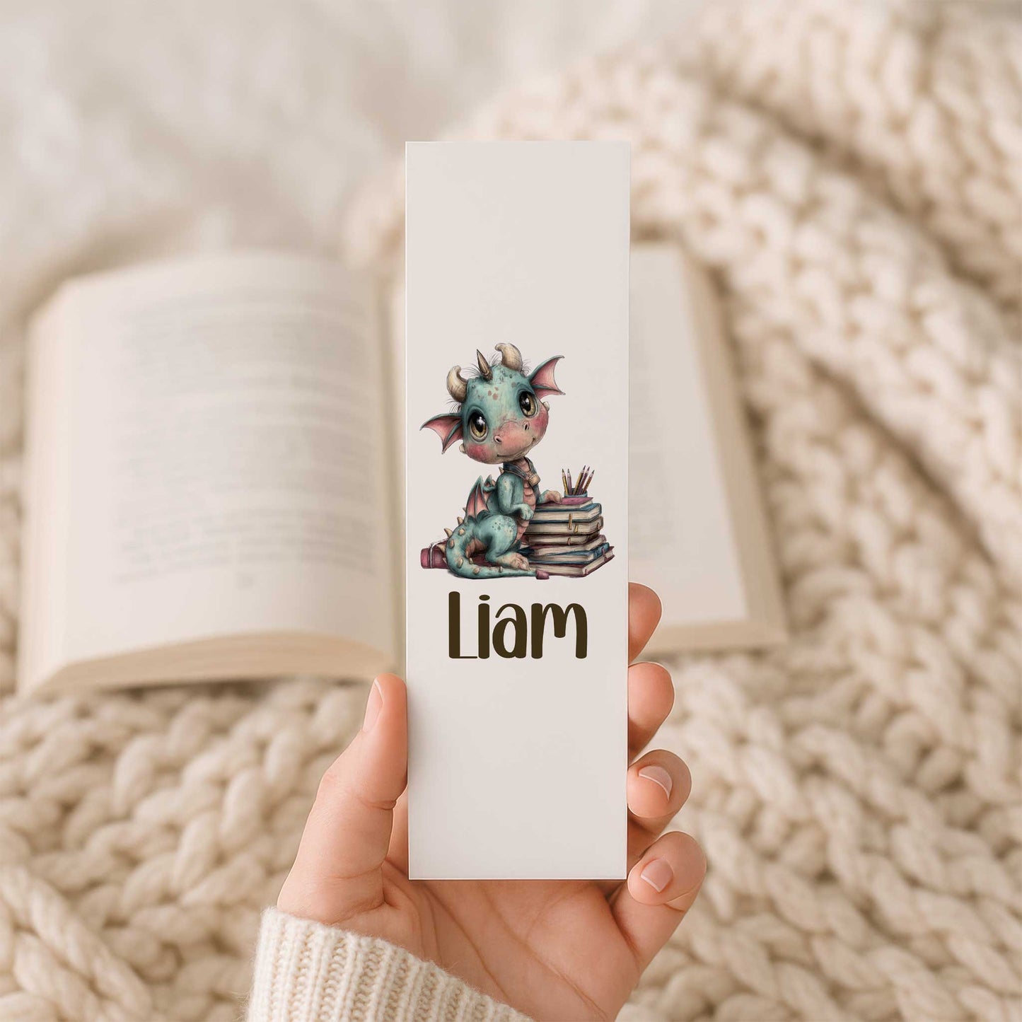 Hand houdt een witte boekenlegger vast met een schattig blauwgroen draakje op een stapel boeken, kleurpotloden en de naam Liam. Op de achtergrond ligt een open boek op een beige gebreide deken.