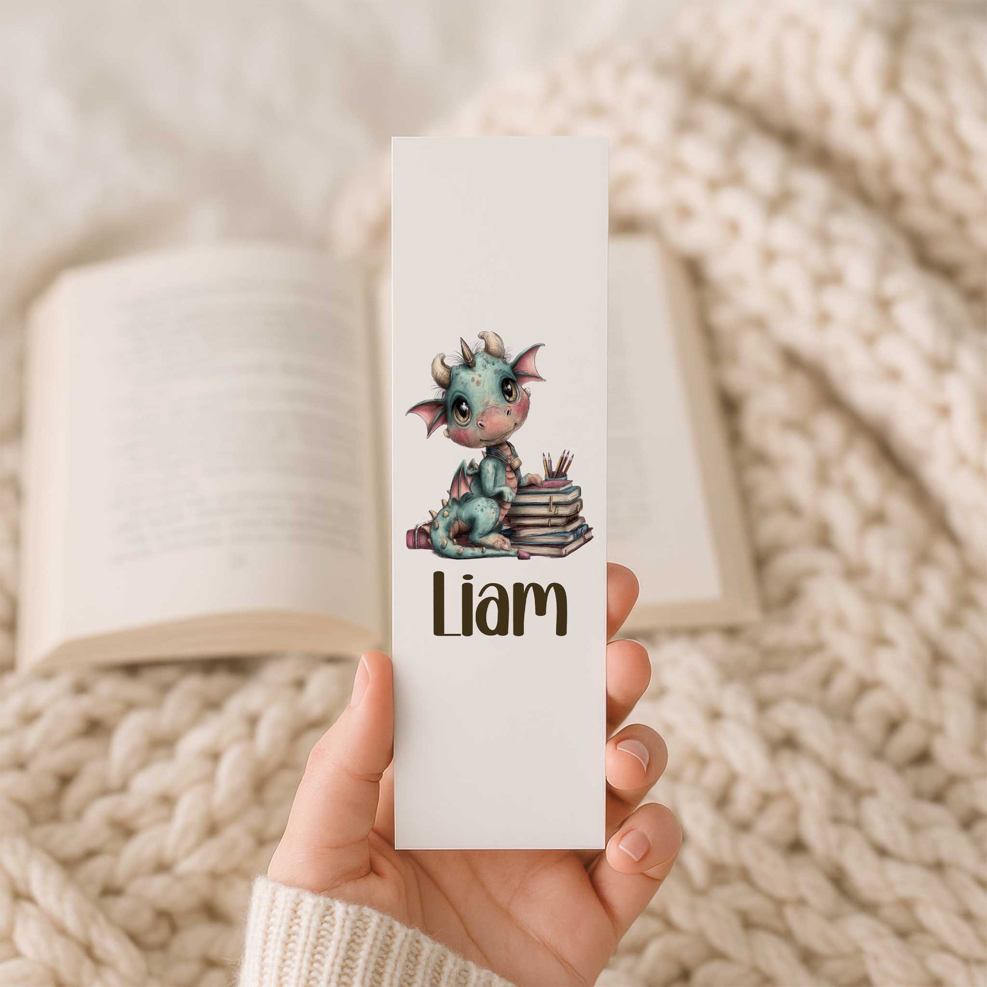 Hand houdt een witte boekenlegger vast met een schattig blauwgroen draakje op een stapel boeken, kleurpotloden en de naam Liam. Op de achtergrond ligt een open boek op een beige gebreide deken.