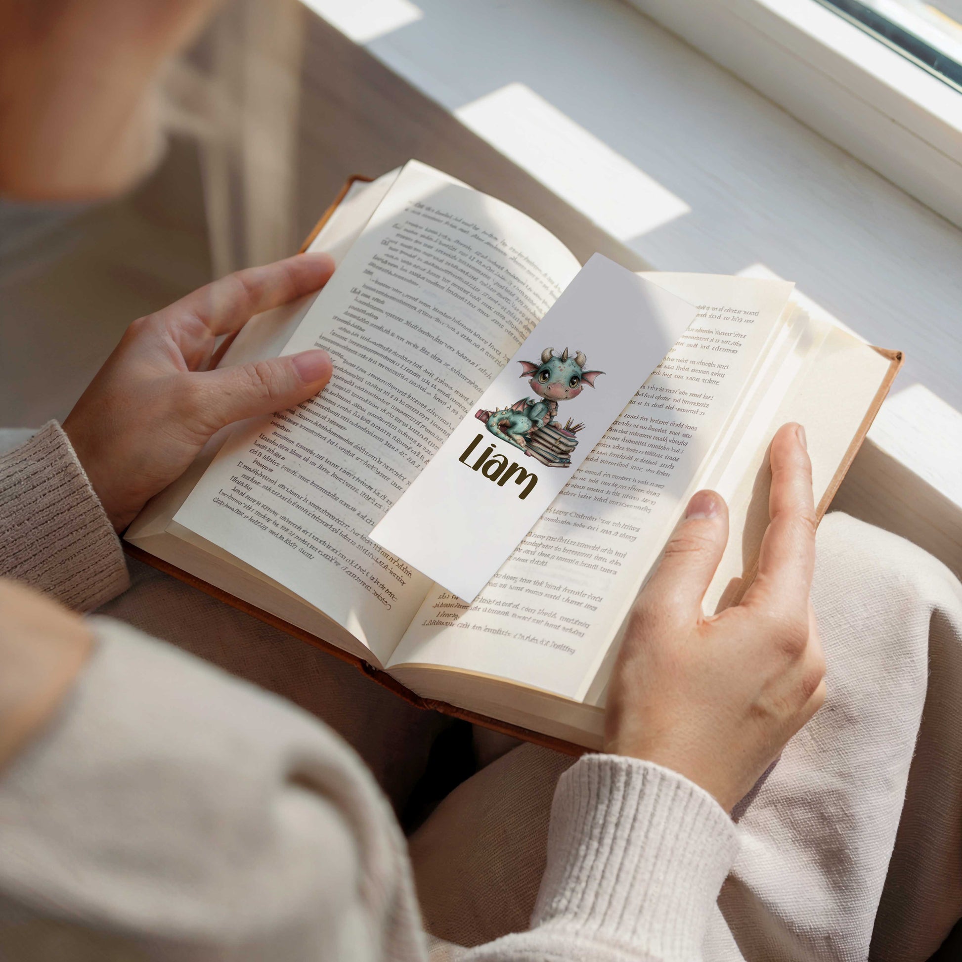 Persoon leest een open boek waarin een witte boekenlegger ligt met een schattig blauwgroen draakje op een stapel boeken en de naam Liam.