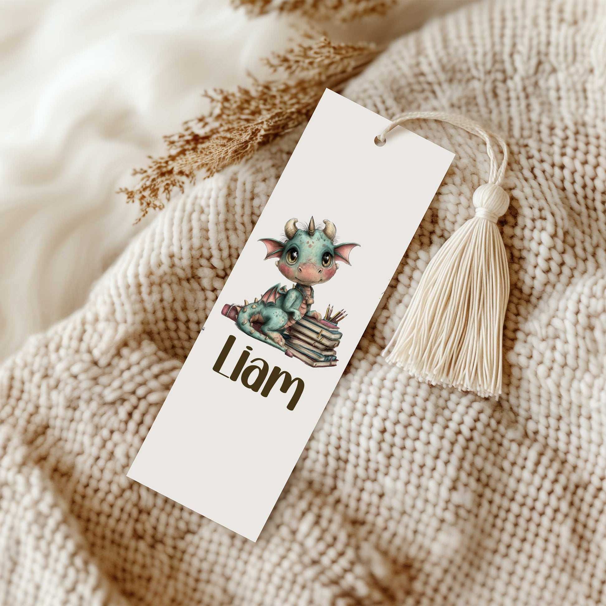 Witte boekenlegger met een schattig blauwgroen draakje op een stapel boeken en de naam Liam. De boekenlegger ligt op een beige gebreide deken en is voorzien van een crèmekleurig kwastje.