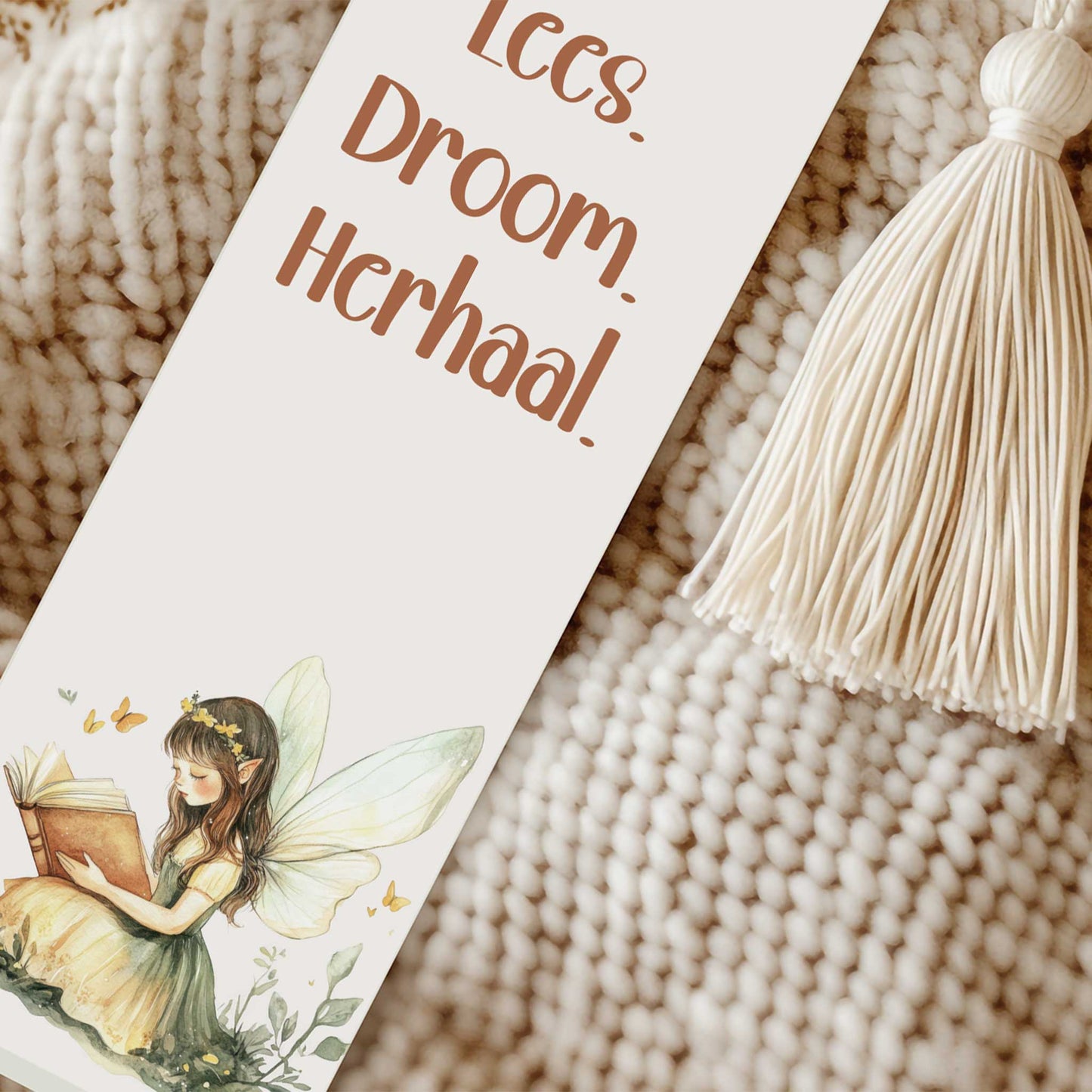 Close-up van een boekenlegger met de tekst "Lees. Droom. Herhaal." naast een crèmekleurig kwastje op een gebreide beige ondergrond.