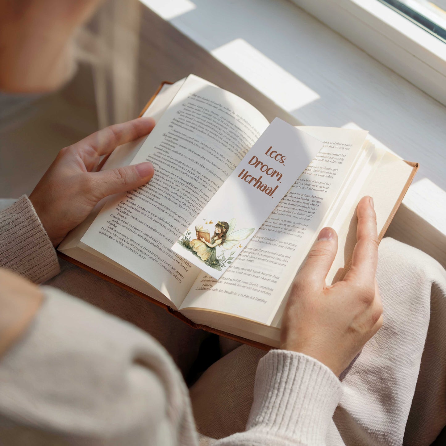Persoon houdt een open boek vast bij een raam met een boekenlegger waarop de tekst "Lees. Droom. Herhaal." en een illustratie van een lezende fee zichtbaar zijn