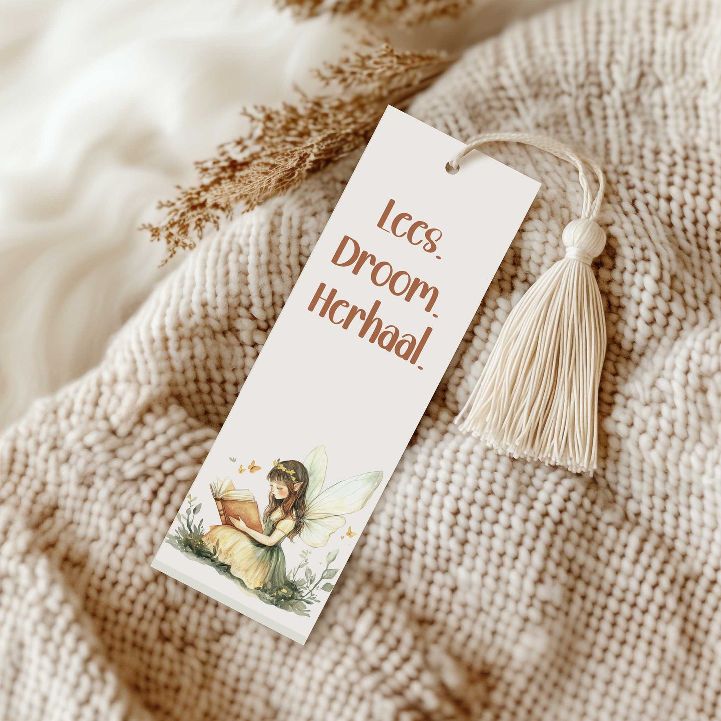 Boekenlegger met crèmekleurig kwastje liggend op een beige gebreide deken naast gedroogde takjes. Op de boekenlegger staat een lezende fee met de tekst "Lees. Droom. Herhaal.".