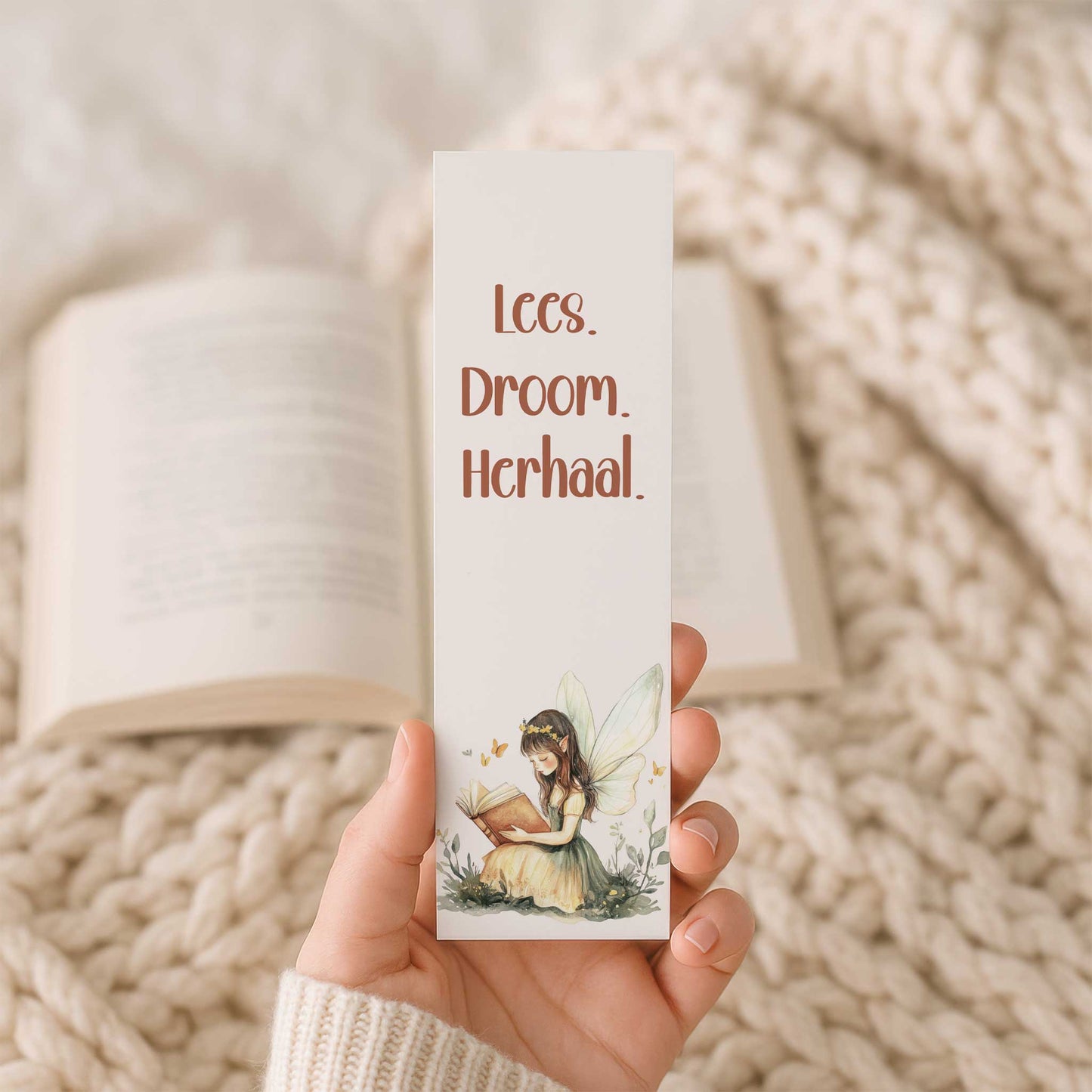Hand houdt een boekenlegger vast voor een open boek op een beige gebreide deken. Op de boekenlegger staat de tekst "Lees. Droom. Herhaal." boven een lezende fee.