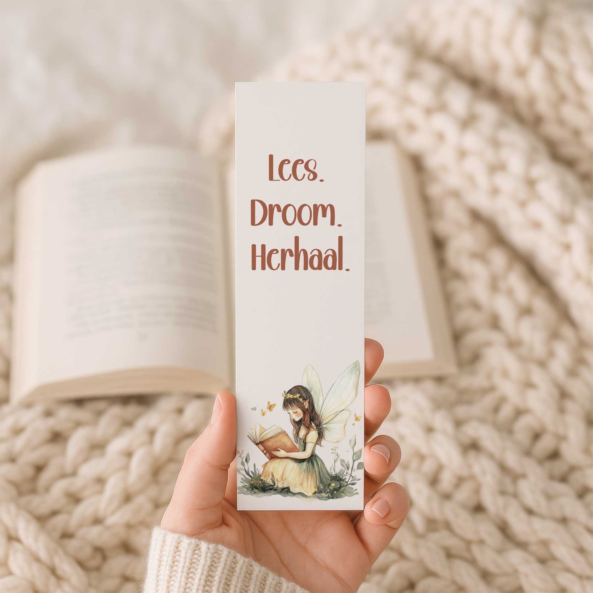 Hand houdt een boekenlegger vast voor een open boek op een beige gebreide deken. Op de boekenlegger staat de tekst "Lees. Droom. Herhaal." boven een lezende fee.