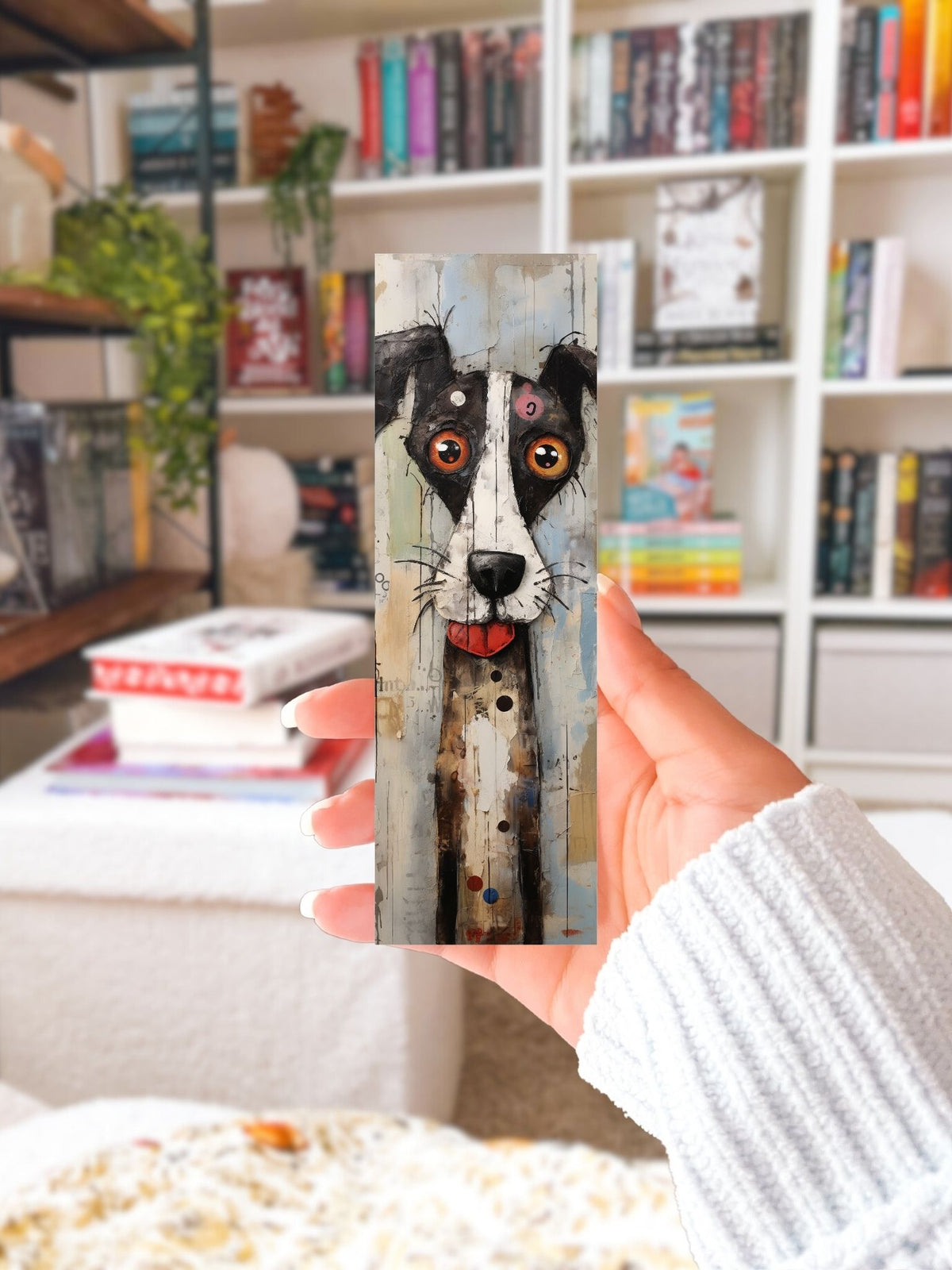 Hand houdt een boekenlegger met langwerpige zwart-witte hond, grote oranje ogen en flapoor voor een boekenkast.