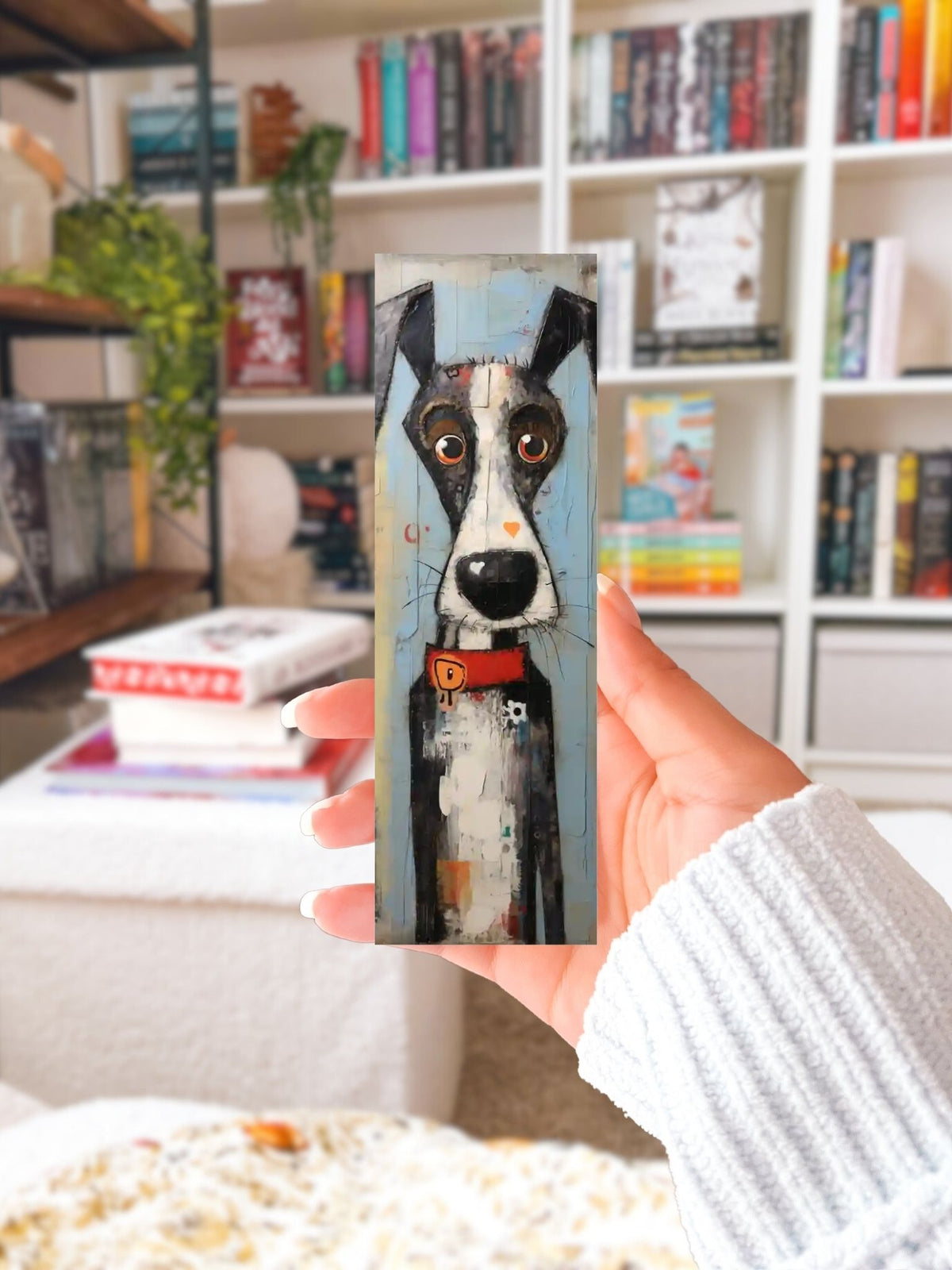 Hand houdt een boekenlegger met zwart-witte hond, grote ogen en rode halsband voor een boekenkast.