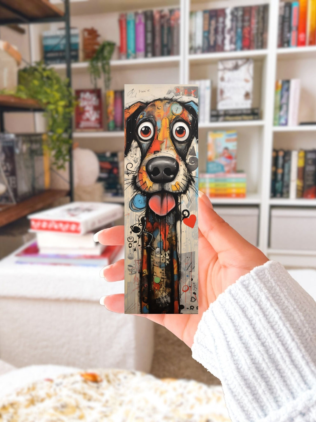 Hand houdt een kleurrijke boekenlegger met grote hondensnoet vast voor een boekenkast met boeken.